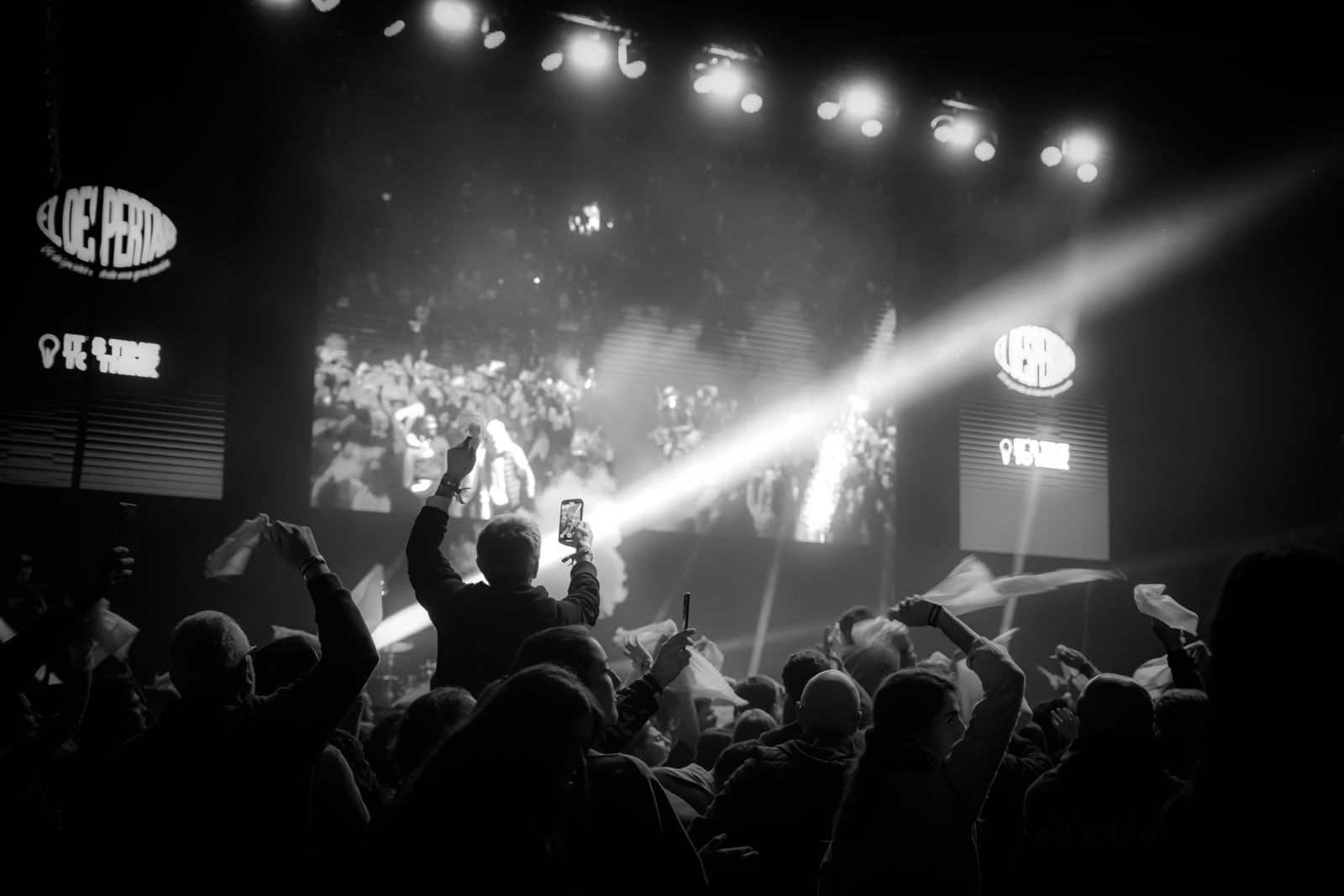 Fotografía en blanco y negro del público frente a la pantalla central y las dos pantallas verticales del escenario.