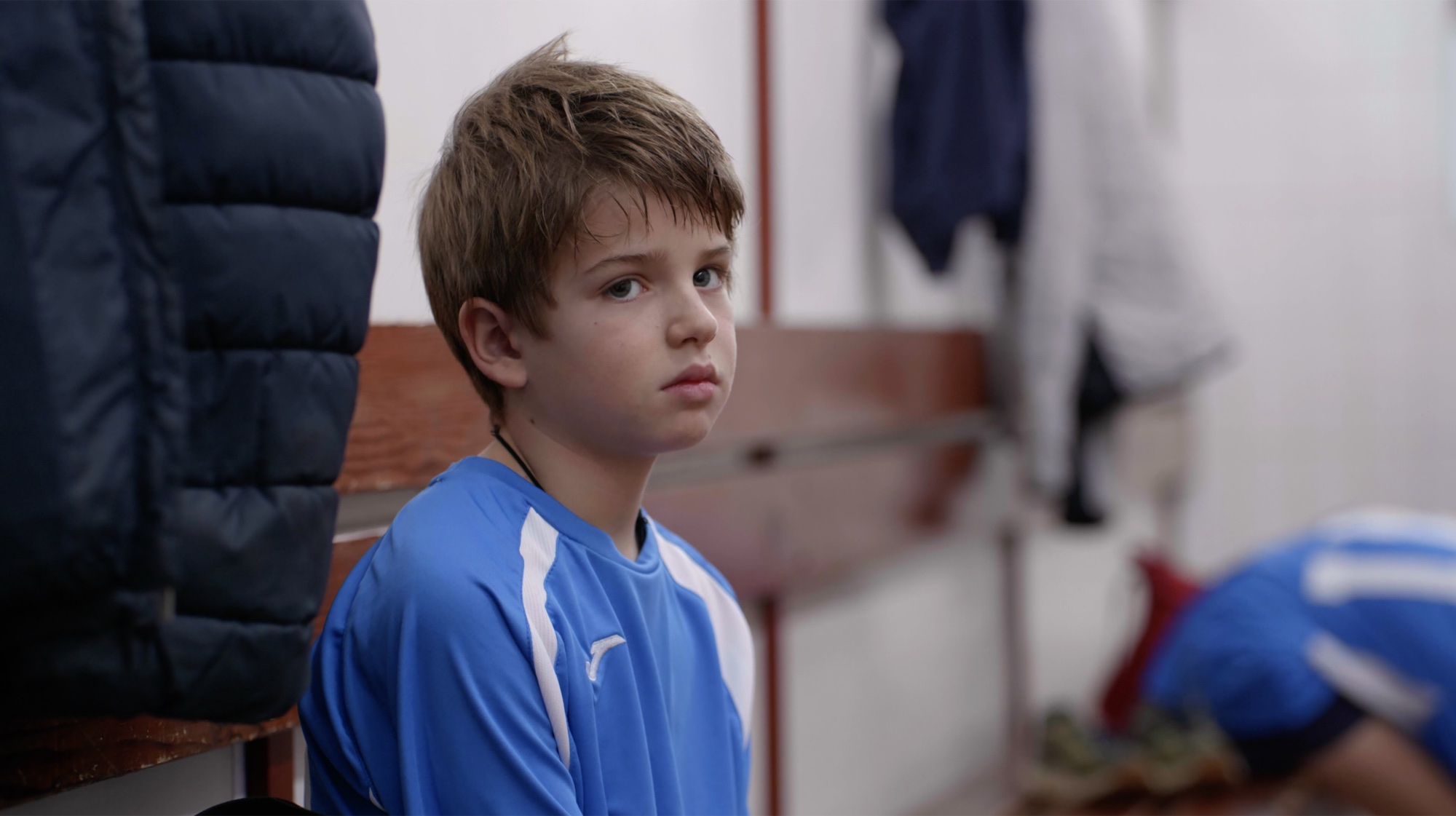 Un niño con la camiseta azul de su equipo mira hacia un adulto desde el banco del vestuario.